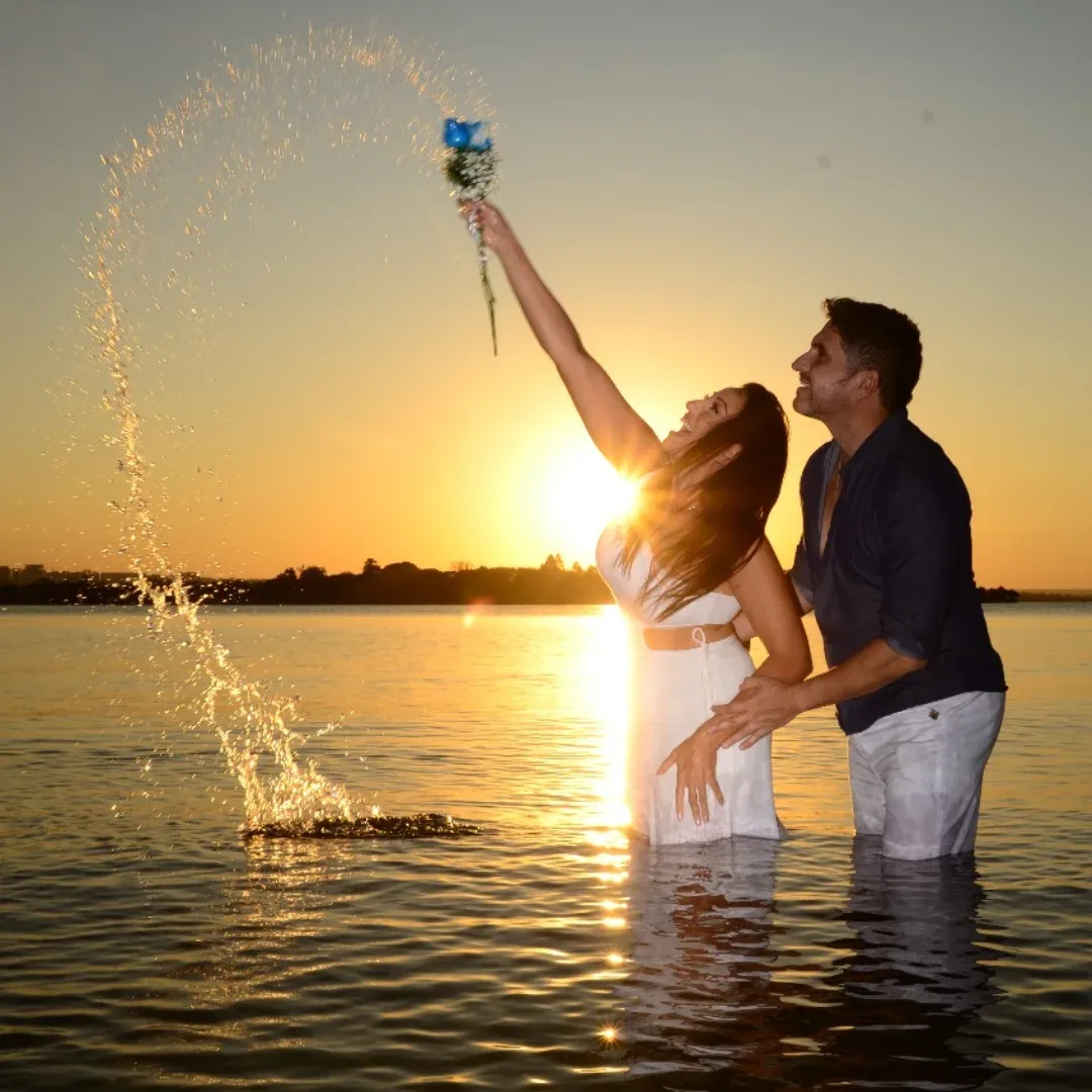 Casal dentro da água ao pôr do sol; a mulher levanta um pequeno buquê e espalha água.