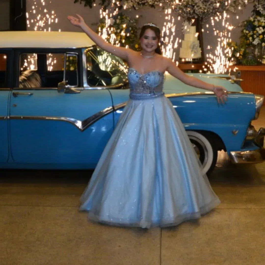 Mulher de vestido azul abre os braços diante de um carro antigo, sob luzes de festa.