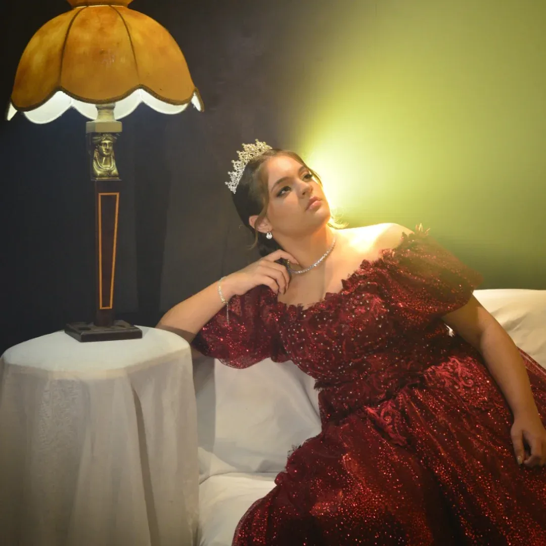 Mulher de vestido vermelho posa sentada junto a um abajur em um interior escuro.