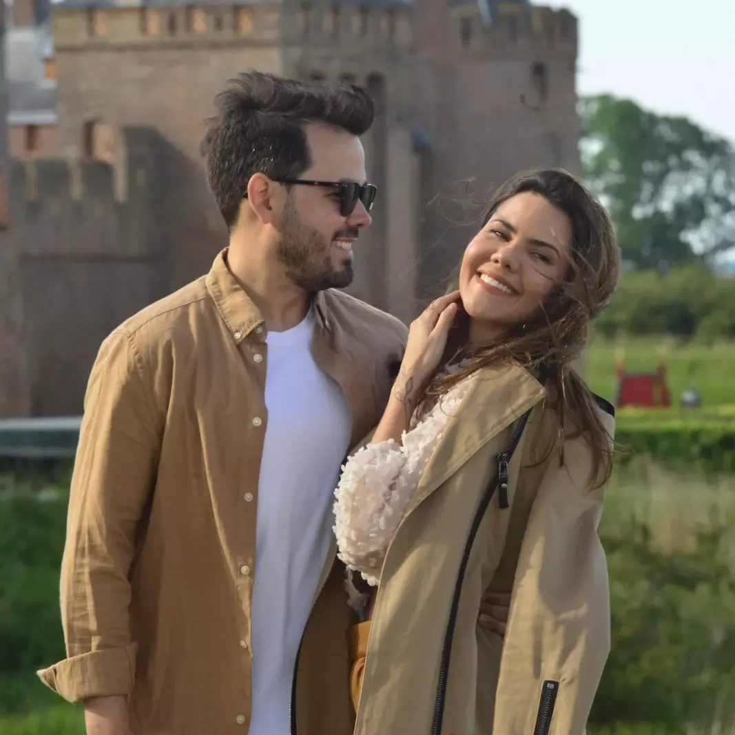 Casal de roupas claras se olha sorrindo diante de uma cerca de madeira em um jardim.