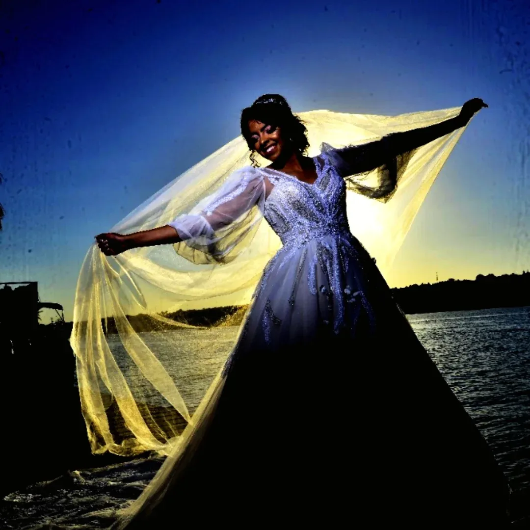 Mulher de vestido azul segura um tecido iluminado em amarelo junto à água ao entardecer.
