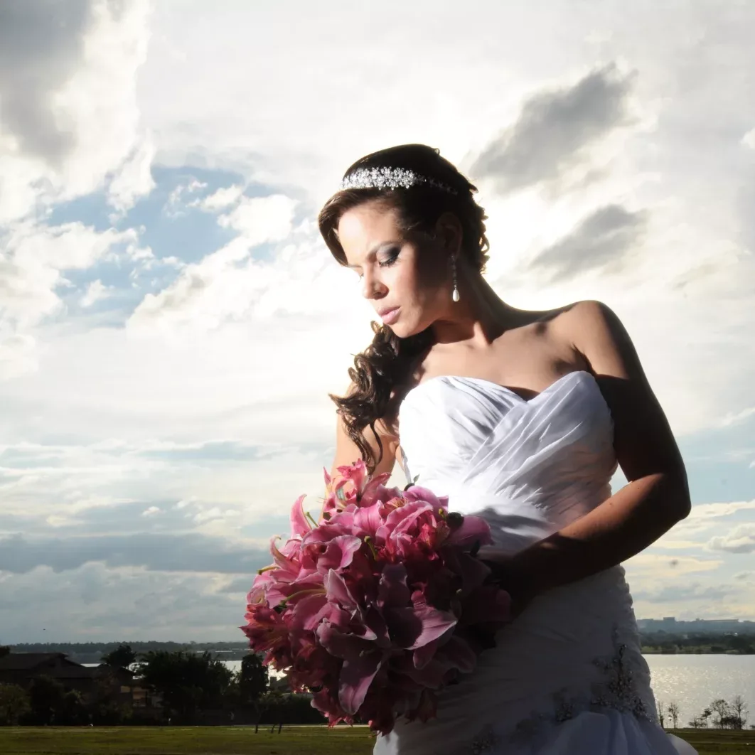 Noiva olha para um buquê rosa em um jardim, sob céu nublado.