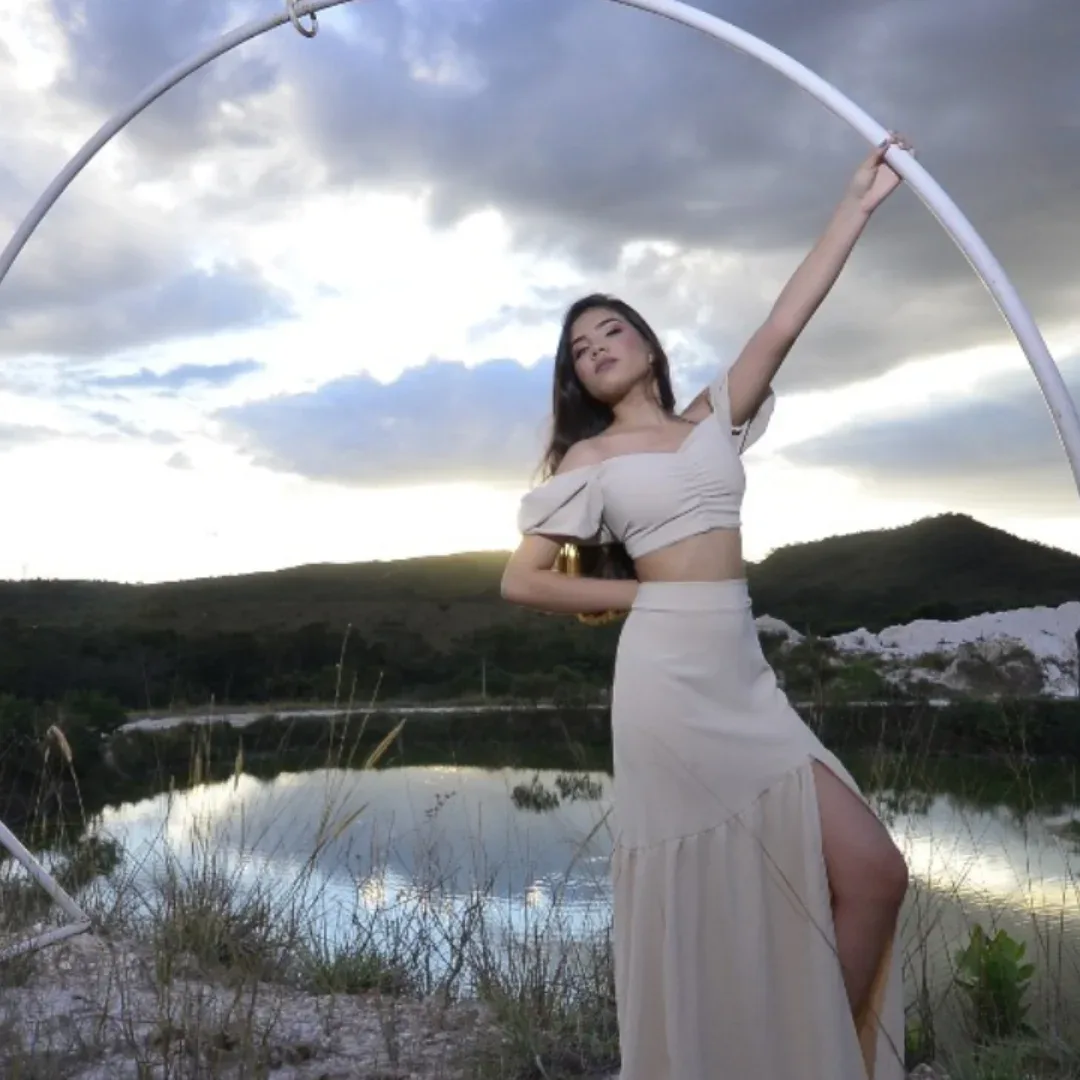 Mulher de vestido claro segura um arco branco diante de um lago e céu nublado.